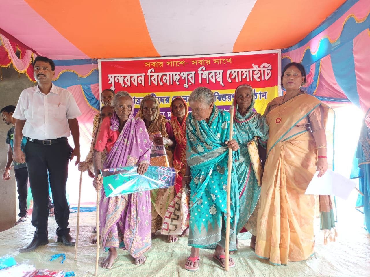 Elderly women receiving saris from society members under a decorated tent